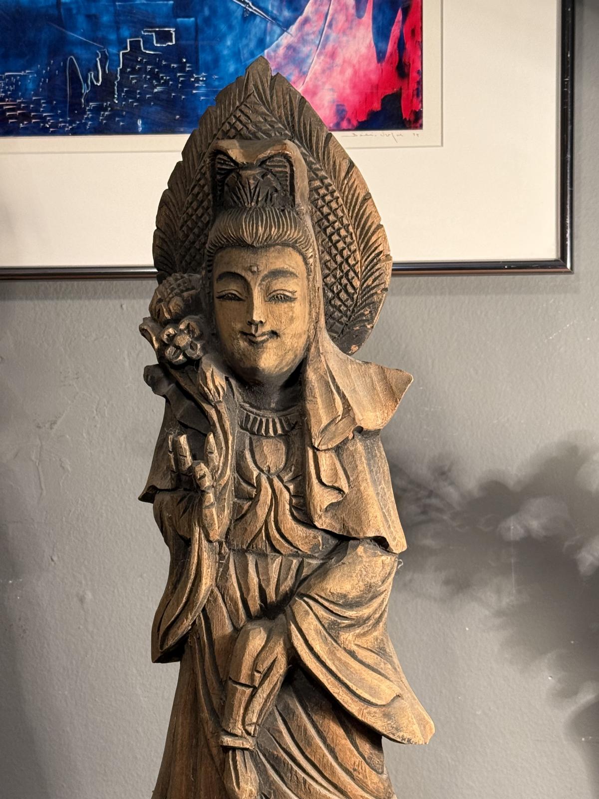 Statuetă orientală din lemn sculptat, cu veșminte drapate și patină brună. Înălțime 55 cm. Prezintă fisuri și lipsuri de material la bază.