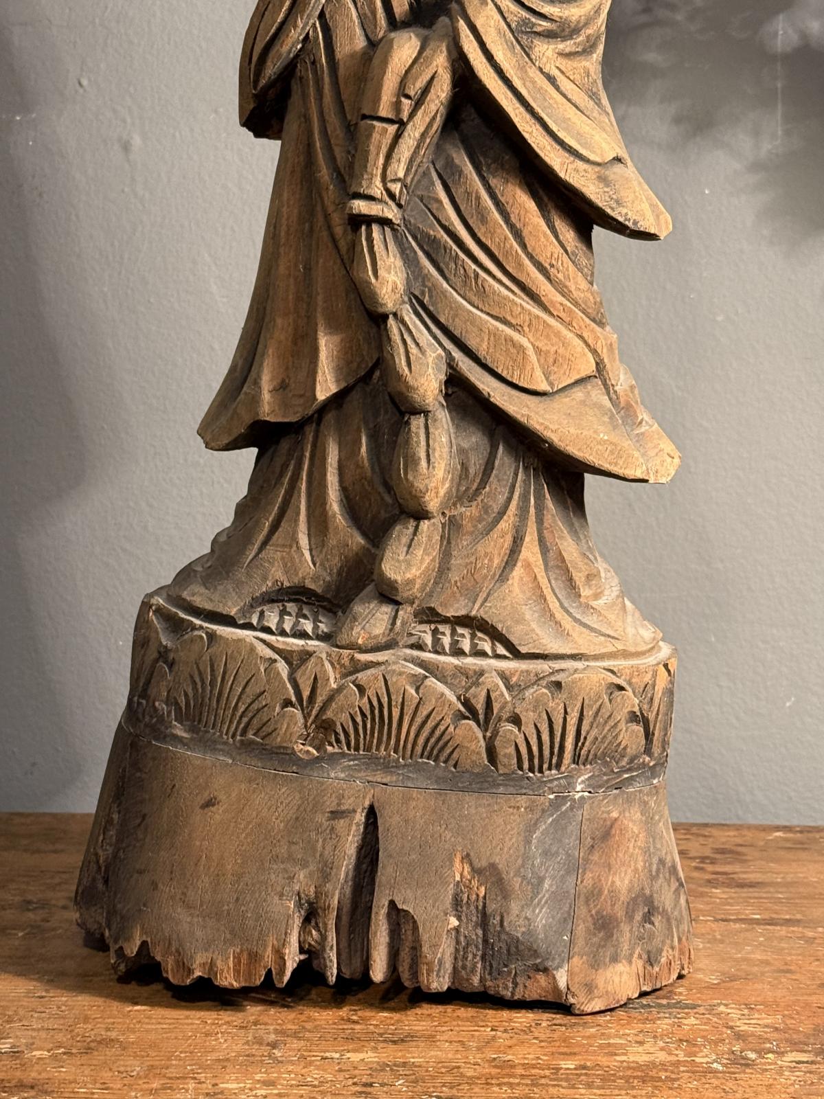 Statuetă orientală din lemn sculptat, cu veșminte drapate și patină brună. Înălțime 55 cm. Prezintă fisuri și lipsuri de material la bază.